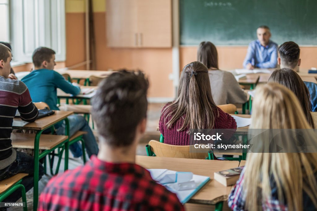 Back view of large group of students sitting in the classroom and listening to their teacher. Focus is on girl in purple sweater.